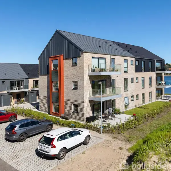 En moderne tre-etagers bygning med altaner, stående tæt på en sø. Der er flere parkerede biler foran bygningen, og omgivelserne er frodige med grønne arealer under en klar blå himmel.