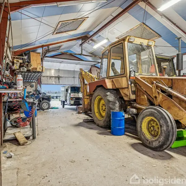 En gammel, gul rendegraver står parkeret i en stor garage eller værksted. Værkstedet indeholder hylder med værktøj og andre biler i baggrunden.