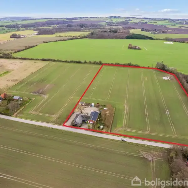 Indhegnet mark med bygninger langs en vej, omgivet af åbne marker og spredte træer, i en landlig og kuperet landskabskontekst.