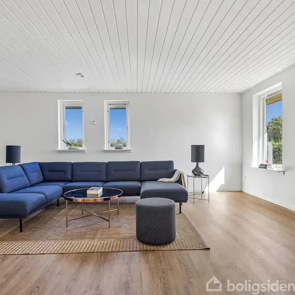 Spisebord med fire stole står i et moderne køkkenalrum med hvidmalede vægge og trælaminatgulv. Over bordet hænger en flettet lampe. Køkkenet har mørke skabe og to vinduer.
