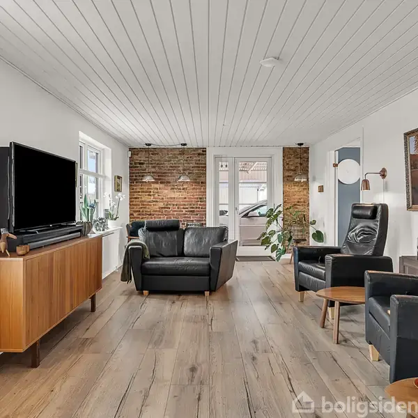 Stue med sorte lædersofaer og lænestole arrangeret omkring et tv på et træskab. Murstensvægge og træloft i baggrunden skaber en hyggelig atmosfære.