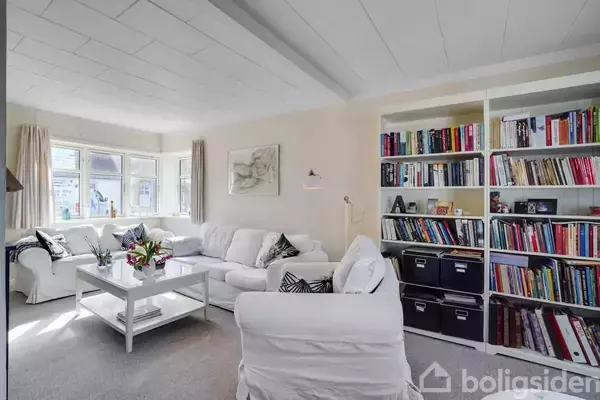 Stue med hvide sofaer omkring et sofabord, blomster som dekoration. Reol fyldt med bøger og opbevaringskasser. Lyse gardiner hænger ved vinduerne, der giver naturligt lys.