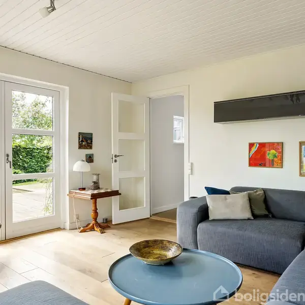 Gråt hjørnesofa står i en lys stue, dekoreret med farverige billeder på væggen. Store glasdøre fører til en terrasse med udsigt til en grøn have med blomstrende rhododendron.