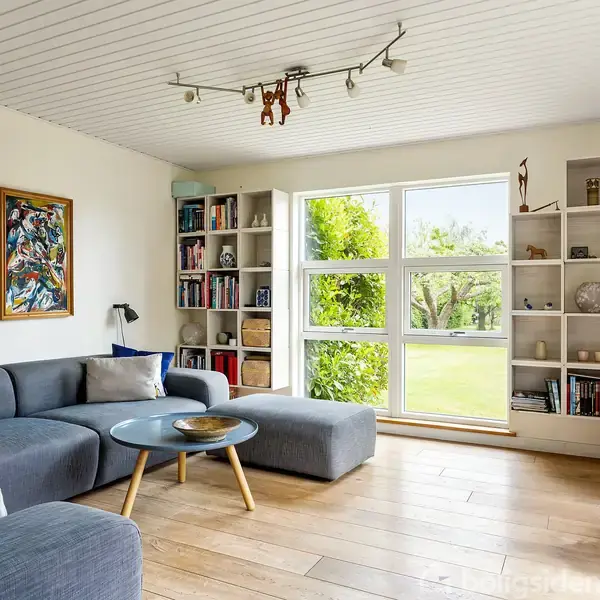 En grå sofa står i en lys stue med parketgulv. En rund glasbord står foran sofaen. Store vinduer viser en grøn have. Bøger og dekorationer fylder hylderne.