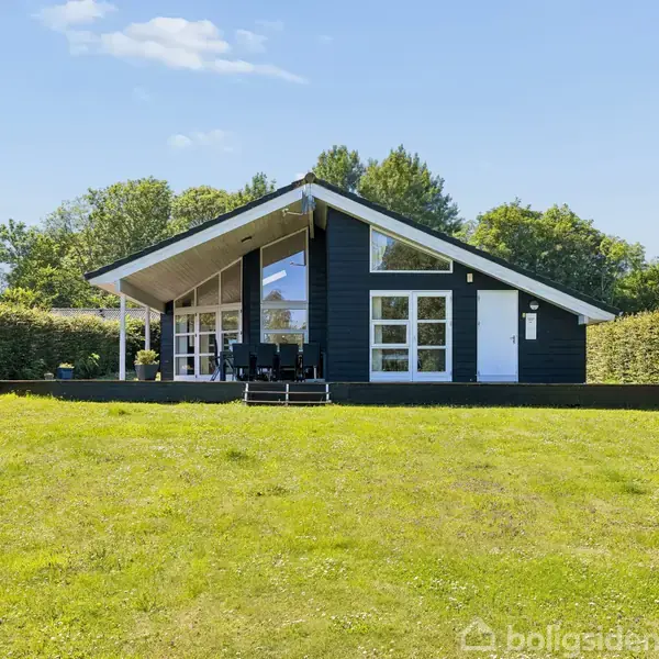 Et moderne sort sommerhus står stille i en velplejet have med grøn græsplæne, omgivet af hække og træer. Terrasse med havemøbler foran huset. Ingen tekst til stede.