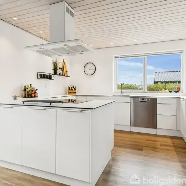 Moderne køkken, hvidt og minimalistisk, med et centralt køkkenø med kogeplade. Store vinduer giver udsigt til en have. Vægge dekoreret med ur og hylder. Gulvet er træ.