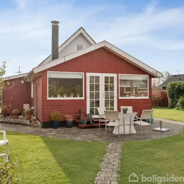 Rødt træhus med skorsten, omgivet af have. Foran huset står bord og stole på flisebelægning. Stien fører fra husdøren til græsplænen. Ingen tekst er til stede.