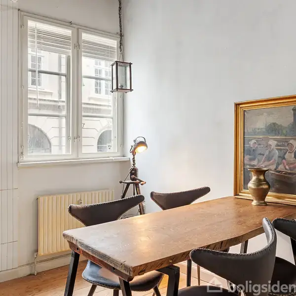 Et træbord omgivet af sorte stole står i et lyst rum. En bordlampe og maleri dekorerer væggen, mens store vinduer lader dagslys strømme ind.