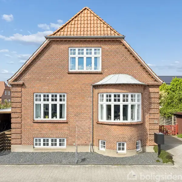 Rødstenshus med rødt tegltag står på en asfaltvej. Huset har hvide vinduer og en rund karnap. Til højre er carport med træhegn, omgivet af buske og træer.