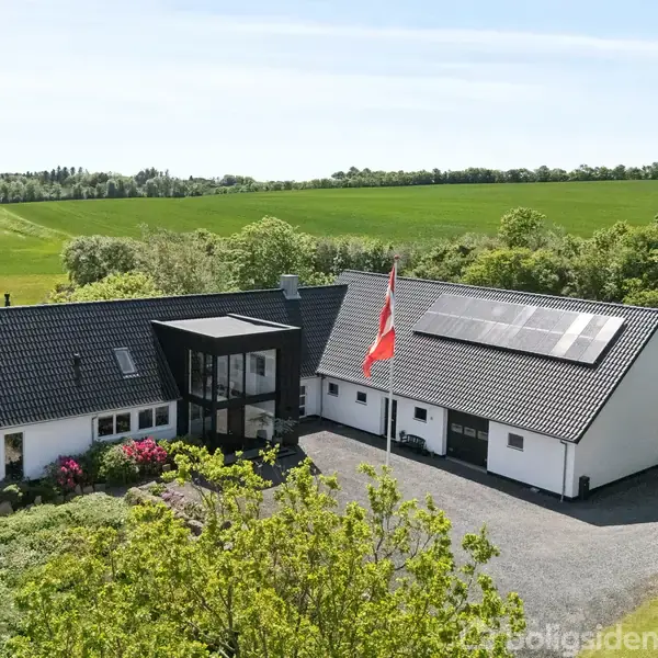 Et moderne, hvidt hus med sorte tage og integrerede solpaneler står foran en flagstang. Det ligger i en landlig scene med store grønne marker og træer omkring.