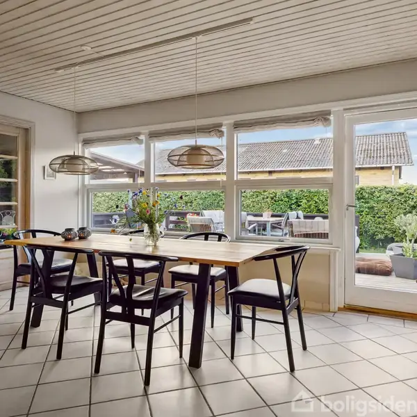 Et træbord med sorte stole står i et lyst spisekøkken. To lamper hænger over bordet. Store vinduer og en glasdør viser udsigt til en terrasse og hæk.