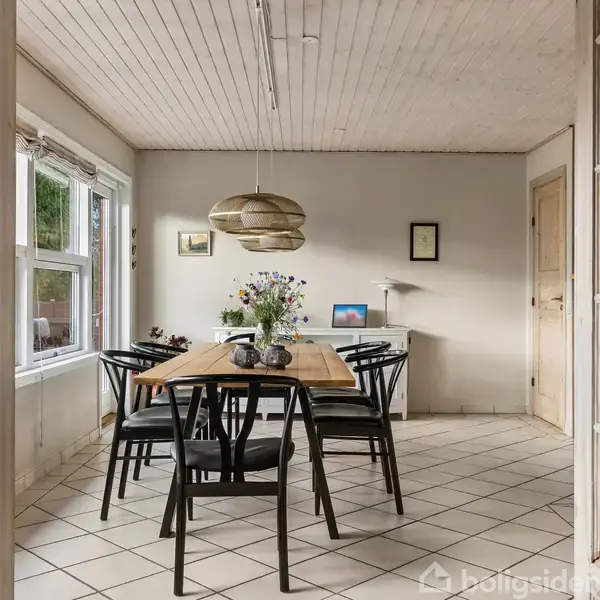 Spisebord med sorte stole står i en spisestue med lyse fliser og træloft. Vaser med blomster pryder bordet. Et skab og en lampe ses i baggrunden.