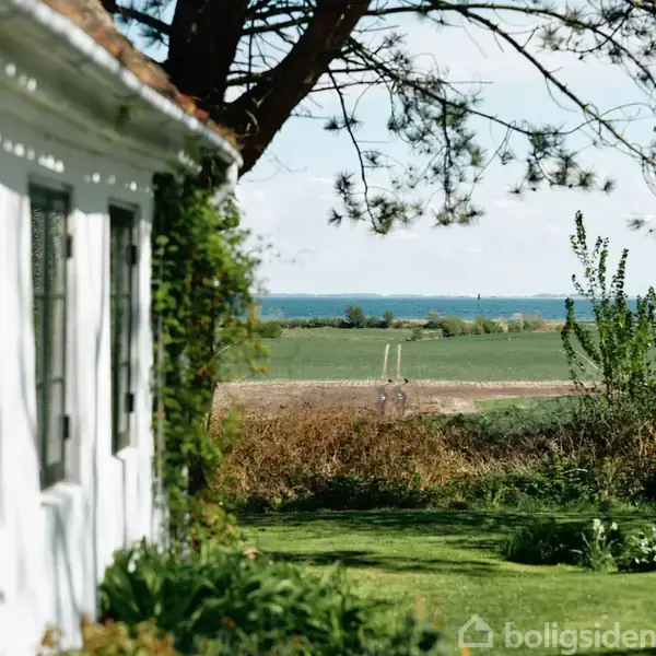 Et hus ses delvist til venstre, med grønt løv, set fra siden. I baggrunden breder marker og hav sig ud under en klar blå himmel.