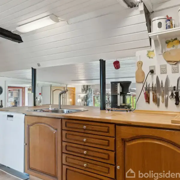 Køkkenbord i træ med indbygget vask og opvaskemaskine, omgivet af køkkenredskaber og knive hængende på væggen. Lys trælignende loft og hylder med opbevaringskrukker i baggrunden.