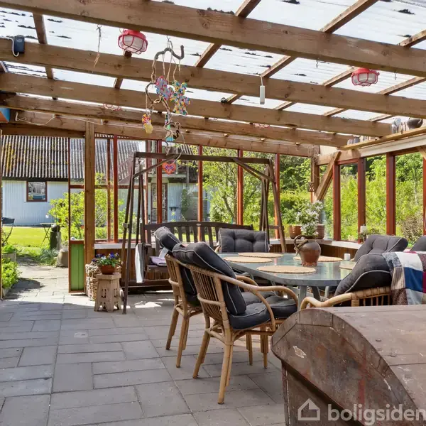 En overdækket terrasse med træmøbler og bløde puder står på flisebelægning. En stor, rund glasbord er omringet af stole, mens farverige dekorationer hænger fra loftet. Grønt haveområde ses udenfor.