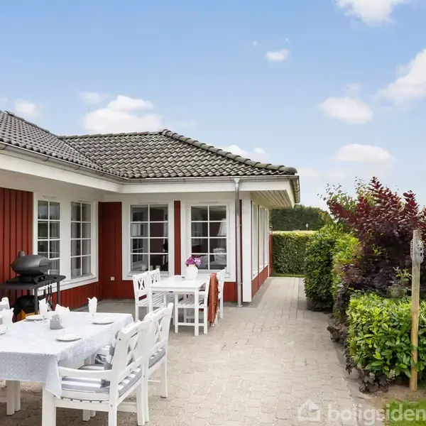 Et rødmalet hus med hvide vinduesrammer har en terrasse med hvide møbler og grill. Omgivet af velplejet græsplæne og hæk, under en blå himmel med få skyer.