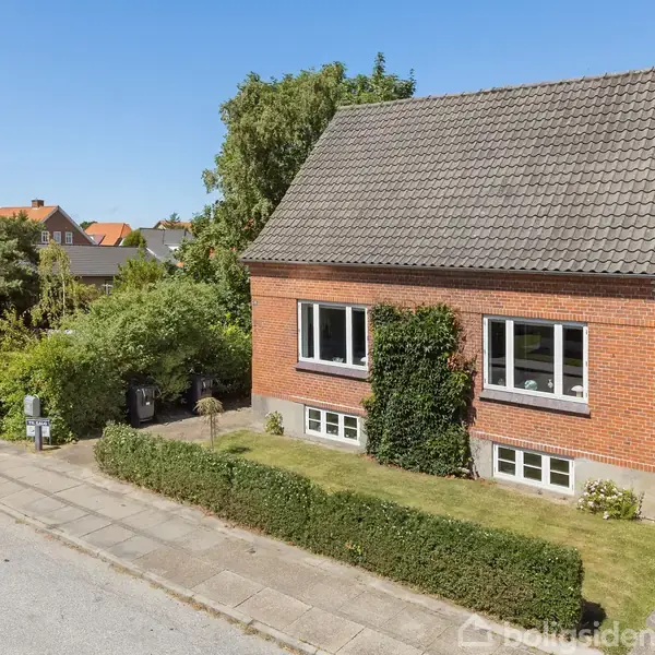 Et rødt murstenshus står stille omgivet af en velplejet have med træer og buske. Huset har et skråt tag og store vinduer. En bil er parkeret ved siden af.