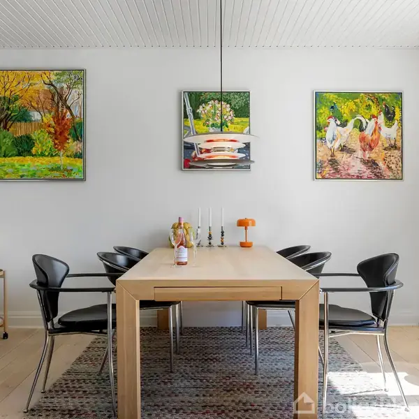 Et spisebord med seks sorte stole står i et lyst rum. På væggen hænger tre farverige malerier. En plante og dekorationer pryder omgivelserne, der er hjemlig og stilfuld.