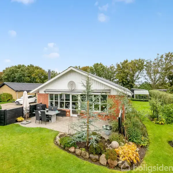 Et rødt murstenshus med store vinduer står i en frodig have. Der er en terrasse med bord og stole i forgrunden, omgivet af velplejede buske og grønt græs.