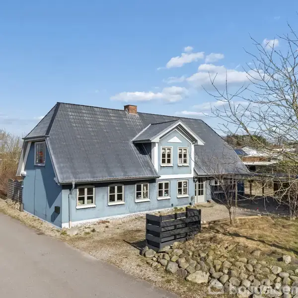 Et blåt to-etagers hus står stille ved siden af en vej. Træer og en have omkranser huset, og en klar blå himmel er i baggrunden.