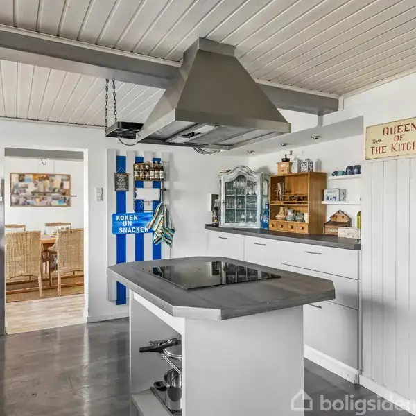 Et moderne køkken med en kogeø i centrum, emhætte over, grøn retro køleskab til højre. Skilt: "QUEENS of the KITCHEN". Indgang fører til spisestue.