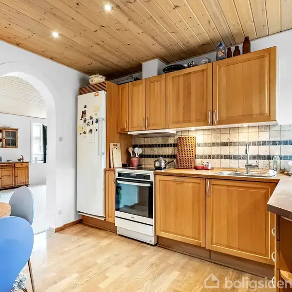 Et køkken med træskabe og indbyggede hvidevarer, herunder en ovn og køleskab. Køkkenet har en farverig flisevæg og træloft. I baggrunden ses en spisebordsstol og en buet indgang.