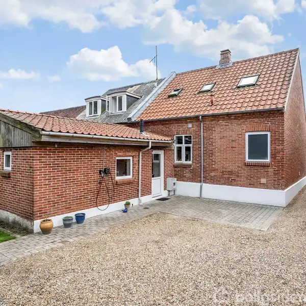 Rødt murstenshus med skråt tag, står stille. omgivet af grus og flisebelagt gårdsplads. En lille tilbygning har nogle plantekrukker ved siden. Klar himmel med skyer i baggrunden.