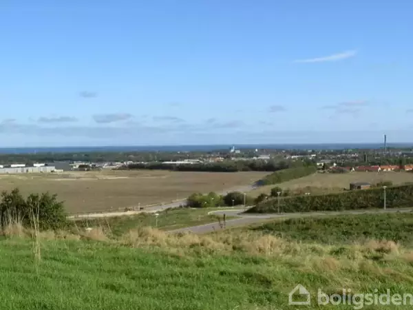 Græsbeklædt bakkelandskab strækker sig mod en by i horisonten, indrammet af marker og spredte vejstrukturer under en klar blå himmel.