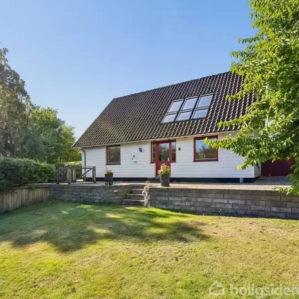 Et hvidt hus med rødt tag står i en solrig have med grønne træer og en græsplæne. Huset har store vinduer og en terrasse.