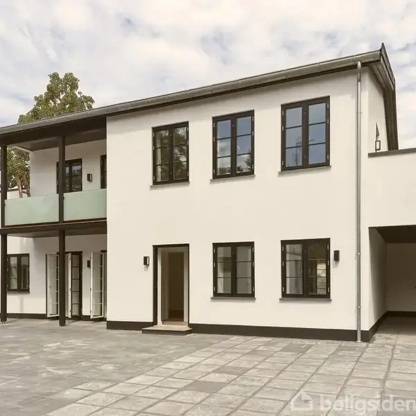 To-etagers moderne hus, står med hvid facade og sorte vinduer. Huset har en overdækket balkon og en stor indkørsel. Det er beliggende i et åbent, flisebelagt område med få træer.