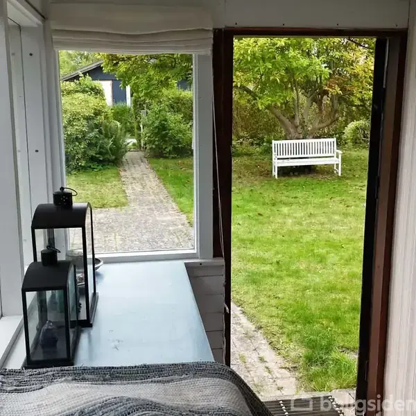 En bænk står i en have, omgivet af grønne buske og træer. Indendørs er der to lanterner på en overflade ved et vindue, der åbner ud mod haven.