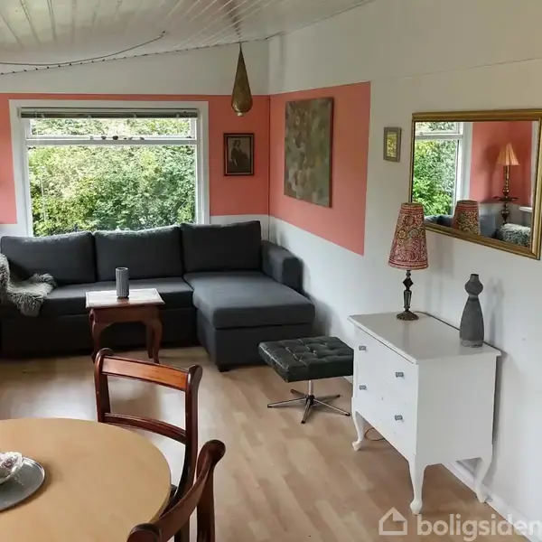 Stue med hjørnesofa foran vindue, dækker udsigt til grønne buske. Rundt spisebord med stole i forgrunden, hvidt konsolbord med lampe og spejl på væggen.