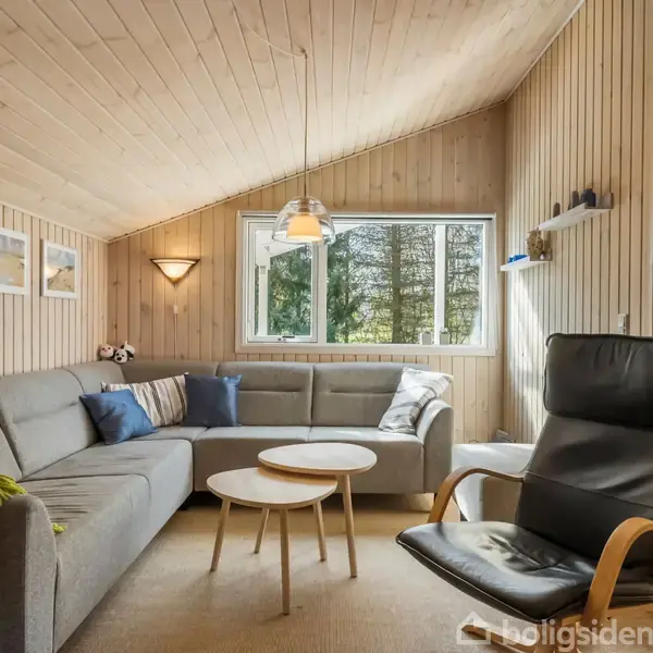 Grå hjørnesofa med farverige puder står i et lyst, træpanelbeklædt rum. Rundt sofabord foran, lænestol til højre. Vindue viser træer udenfor. Lampe hænger fra loftet.