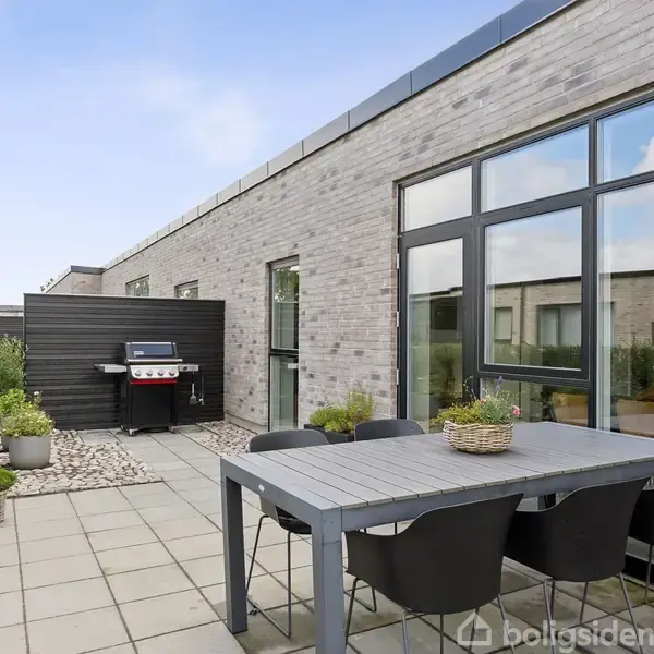 Terrasse med et spisebord og seks sorte stole under en blå himmel. En gasgrill står tæt på, omgivet af forskellige planter i krukker mod en murstensvæg.
