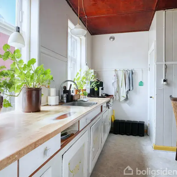 Køkkenskab med træbordplade har planter og køkkenredskaber; det ligger op ad et lyst vindue, mens en kurvestol står ved et spisebord i et hyggeligt køkkenmiljø.