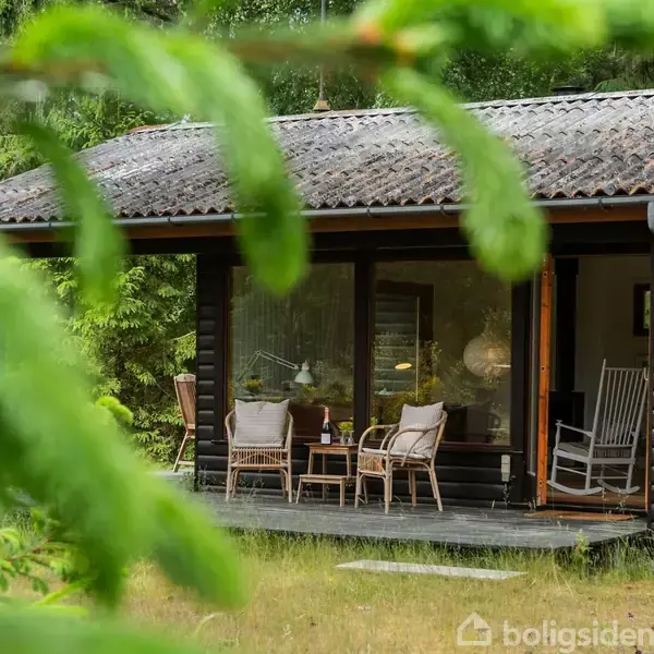 Træhus med overdækket terrasse, flere stole og et bord, omkranset af grønt løv i en skovrig have.