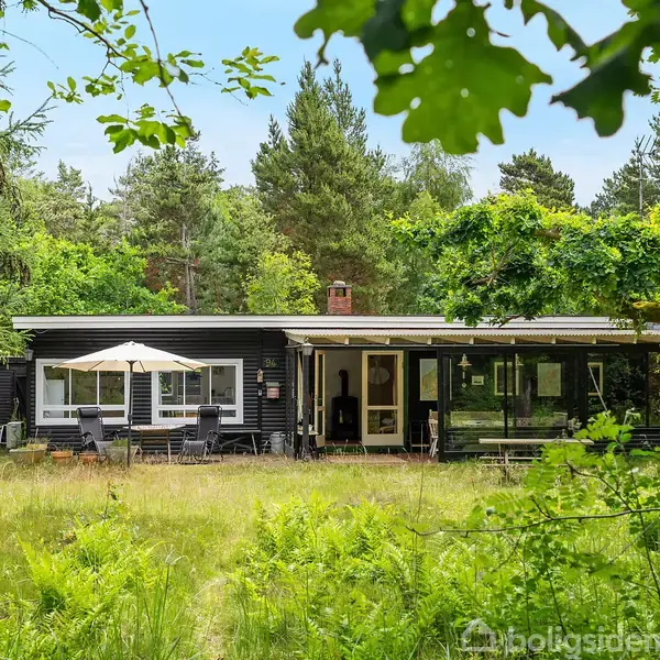 Sommerhus i skoven omgivet af træer. Huset har en mørk facade med store vinduer. En terrasse med parasol er foran huset i en frodig have.