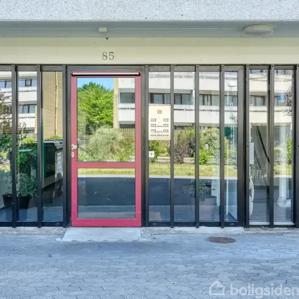 En glasfacade med vinduer omgiver en rød dør, der markerer nummer 85. Baggrunden viser en grøn have og en bygning.