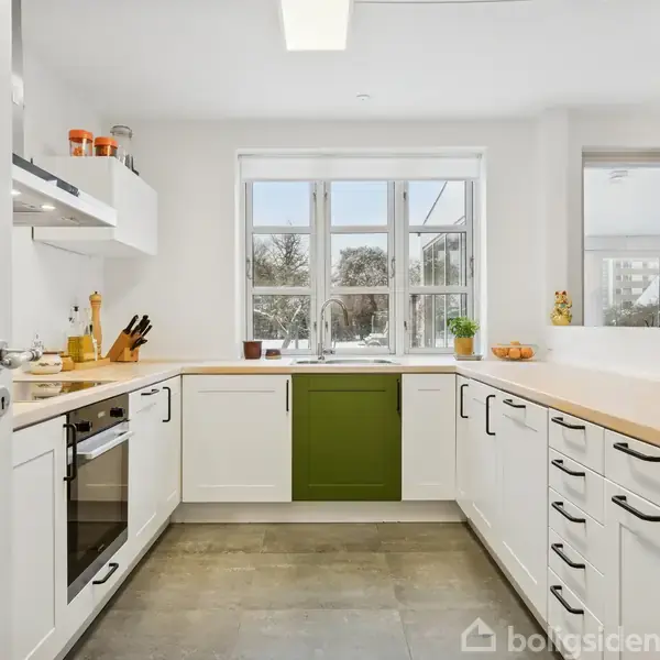 Køkkenet har hvide skabe og en grøn skuffe, bordplade i lyst træ, elektroapparater og vinduer, der giver udsigt til en snedækket have.