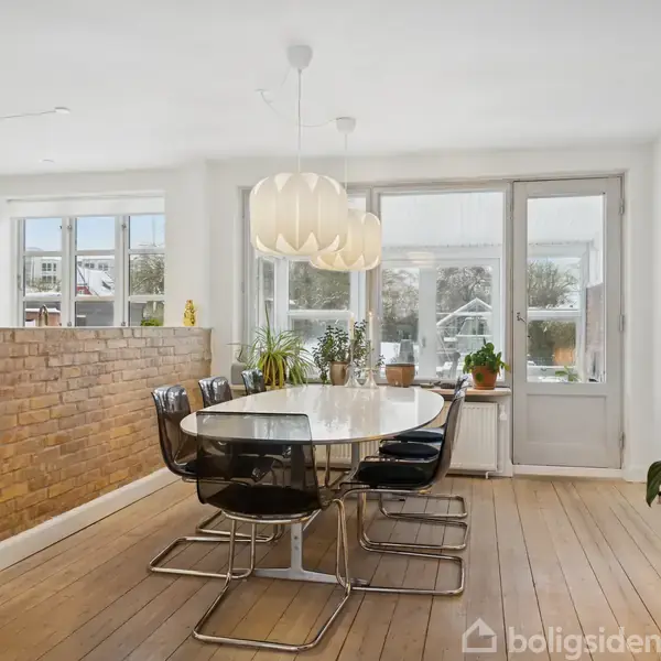 Rundt spisebord med sorte stole står i en lys spisestue. Der hænger en stor lampe over bordet. Rummet har trægulv, murstensvæg og vinduer med udsigt til have.