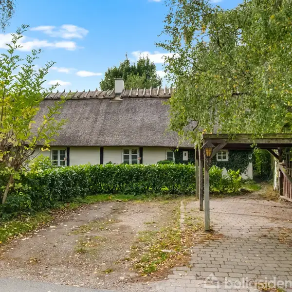 Stråtækt hus står stille bag tæt hæk, omgivet af træer og en brostensbelagt indkørsel. En blå himmel med spredte skyer ses i baggrunden.