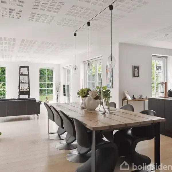 Træbord med sorte stole står i en moderne, lys stue-køkken-kombination. Store vinduer lader naturligt lys ind. En grå sofa ses i opholdsafdelingen. Grøn udsigt udenfor.