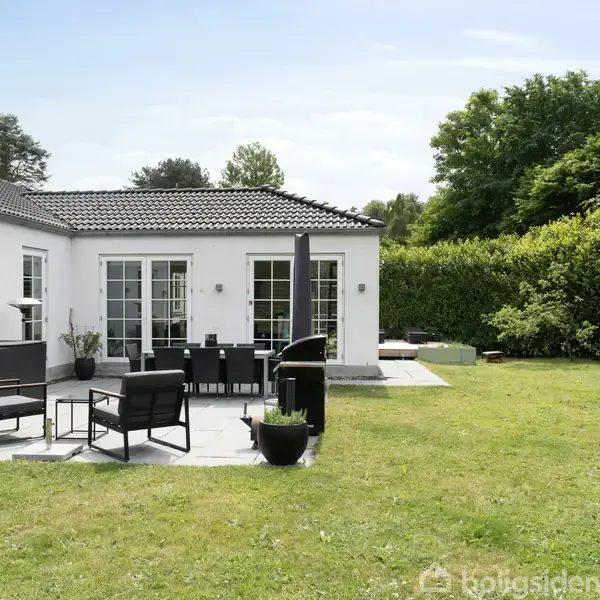 Hus med hvid facade og mørkt tag står i en have. Havemøbler på terrasse, omkranset af græsplæne og buske. Trampolin ses til højre. Grønne træer i baggrunden.