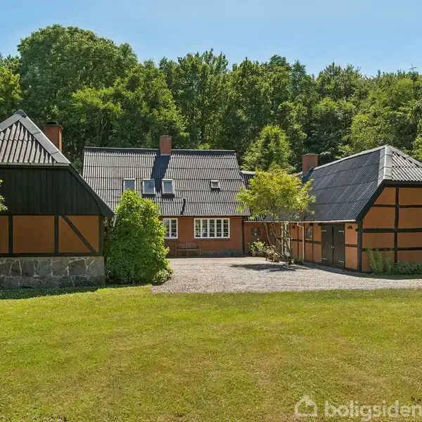 Et traditionelt bindingsværkshus står i en grøn have omgivet af træer. Huset har sort tag og orange vægge med mørke bjælker. Grusgang fører til indgangen.