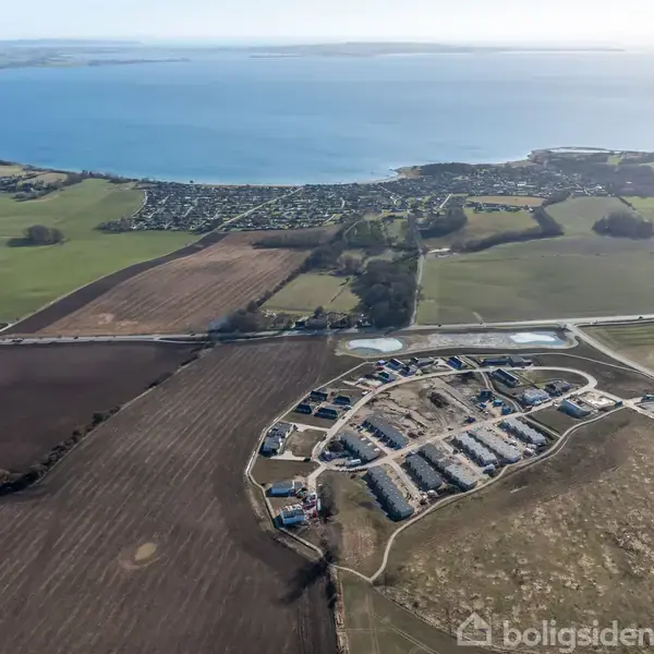 Byområde med nye huse ligger i forgrunden; de er omgivet af marker og veje i landskabet. I baggrunden ses en kystlinje ved havet under en klar himmel.