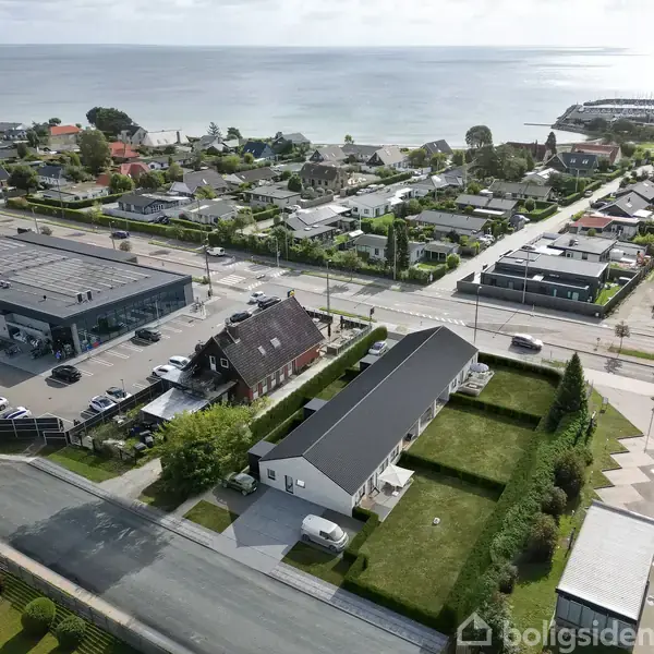 Byområde med huse og forretninger langs hovedvej. To større bygninger og parkeringspladser på venstre side. Havudsigt i baggrunden med kystlinje. Grønt areal og træer omkring.