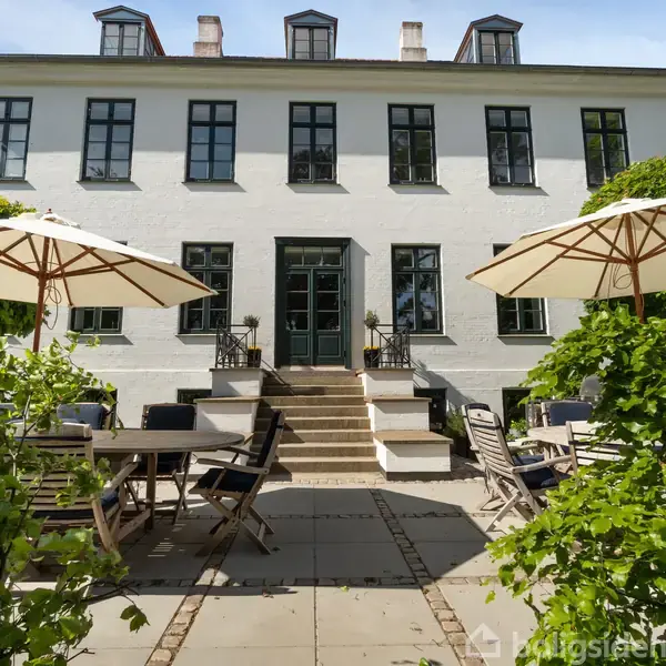 Hvid bygning med sorte vinduesrammer står i baggrunden. Terrassen i forgrunden har havemøbler og parasoller, omkranset af grønne buske. Tre trappetrin leder op til indgangen.