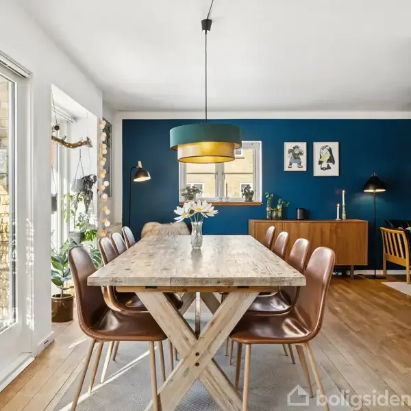 Et træbord med brune læderstole omkring står i en lys spisestue. En moderne lampe hænger over bordet. Blå væg med kunstværker ses i baggrunden.