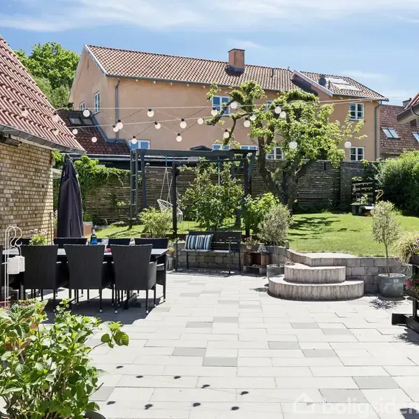 Terrassemiljø med havebord og stole under lyskæder, omgivet af blomsterkrukker. I baggrunden ses en græsplæne med træer, buske og legeredskaber foran en bolig med rødt tag.