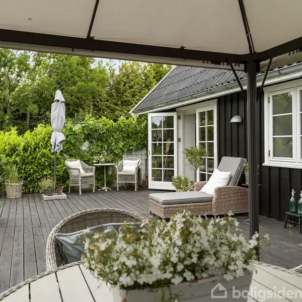 En terrasse med kurvemøbler, stole og liggestol under en parasol. I baggrunden er et sort hus med hvidrammede vinduer, omgivet af grøn beplantning og træer.